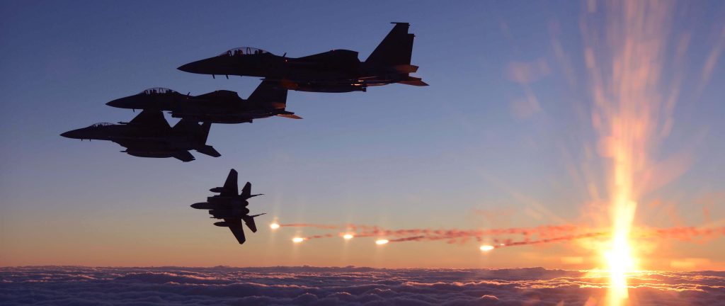 A imagem mostra vários caças militares F-15K Slam Eagle da Coreia do Sul voando em formação ao entardecer, vistos como silhuetas escuras contra o céu. Um dos aviões lança sinais luminosos, que deixam rastros brilhantes no ar. Ao fundo, o sol está se pondo no horizonte, iluminando as nuvens abaixo com tons alaranjados e dourados, criando uma cena dramática e intensa.