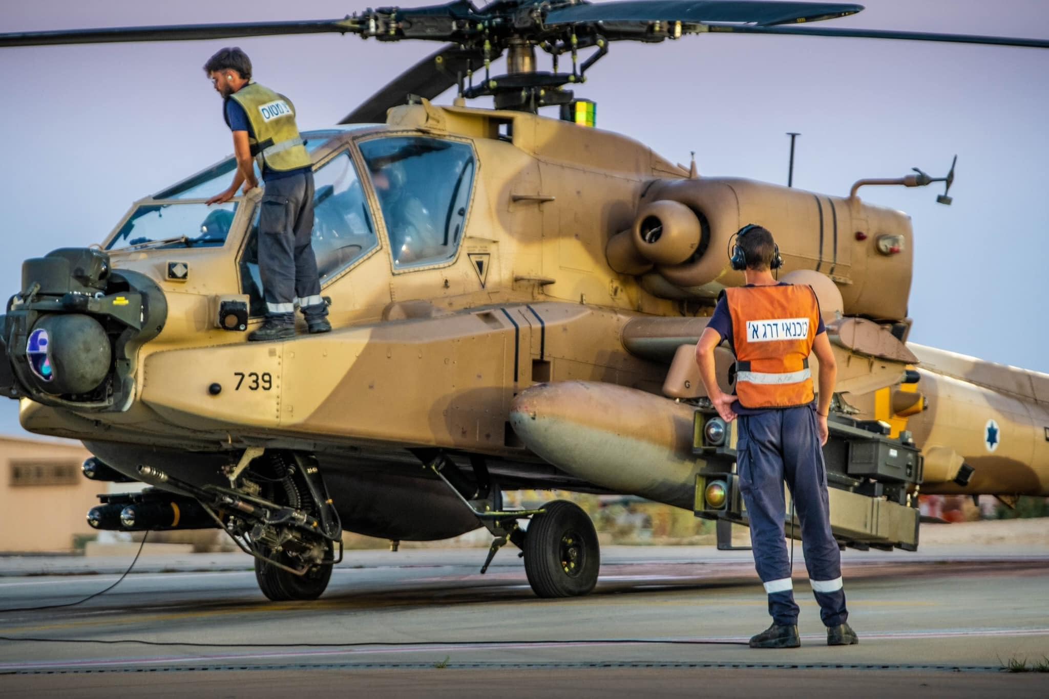 A imagem mostra um helicóptero militar de ataque AH-64 Apache, de cor bege, parado em solo. Dois militares estão ao redor da aeronave: um em pé ao lado, usando colete e fones de ouvido, e outro sobre a fuselagem, próximo ao cockpit, realizando inspeção. O helicóptero tem rotor principal no topo, sensores na parte frontal e aparência robusta, típica de uso militar. A cena sugere preparação ou manutenção antes de uma missão.